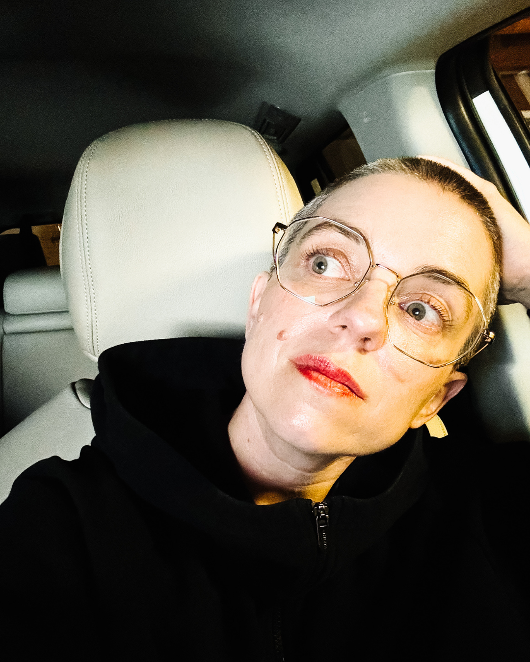 A photo of Meg with a buzzcut, sitting in the front seat of her car in her garage